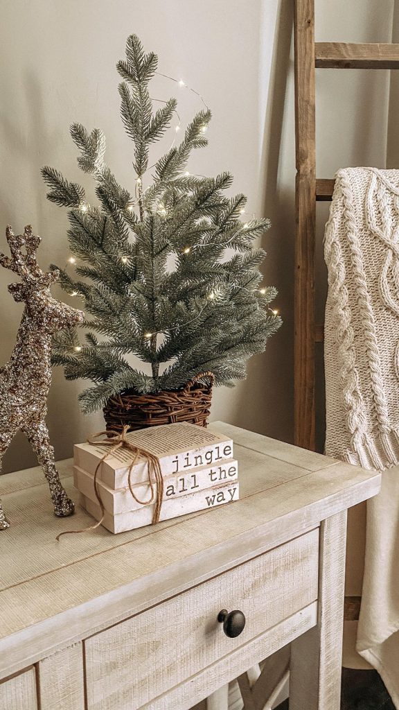 Small artificial Christmas tree on a nightstand beside stacked books labeled with festive holiday terms — minimalist Christmas decor on a budget from By Paige Nicole.